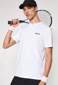 Biele športové tričko s krátkym rukávom, so čiernym logom "BOSS". Hladká tkanina, doplnená bielym náramkom na zápästie a tenisovou raketou.