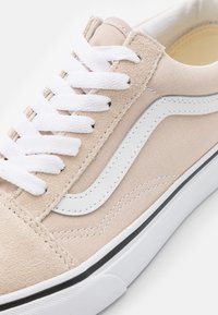 Beige mockaskons med vita snören och en vit sidremsa. Har en svart gummisula och ett strukturerat kanvasövre. Sömnadsdetaljer närvarande.