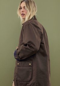 Blond kvinde, der vender sig, iført en brun jakke med ternede manchetter, stående mod en ensfarvet olivengrøn baggrund.
