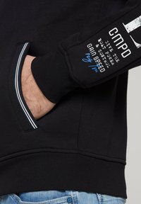 Sudadera negra con un bolsillo tipo canguro, texto blanco y azul en la manga y puños de canalé. Presenta un detalle a rayas en el bolsillo.