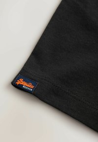 Tricou negru din bumbac cu tiv cusut. Prezintă o etichetă „Superdry” albastră și portocalie pe partea inferioară laterală, evidențiind detaliile brandului.