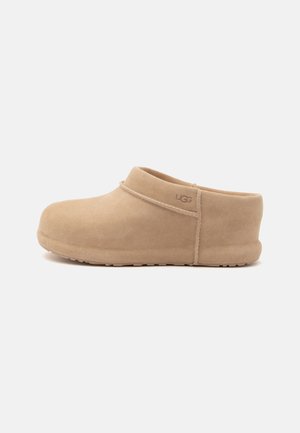 Hellbrauner Veloursleder-Schlupf-Clog mit niedrigem Absatz und UGG-Logo an der Seite, vor schlichten weißem Hintergrund gezeigt.