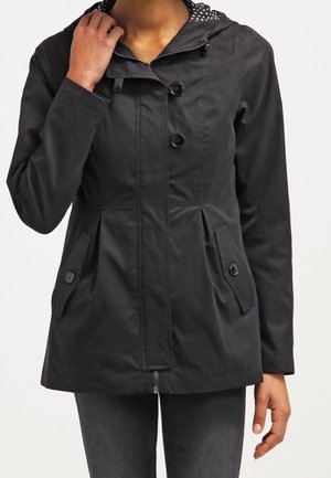 Femme portant une veste noire à boutons avec des poches latérales, tenant le col d'une main, se tenant devant un fond uni.