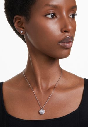 Silberne Kettenhalskette mit einem herzförmigen Anhänger, der mit einer Ansammlung von klaren Steinen verziert ist. Kleine Steckerohrringe mit einem einzelnen Stein. Schwarzes Oberteil.