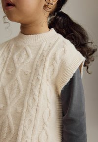 Cremfarbener gestrickter Pullunder mit Zopfmuster, geripptem Halsausschnitt und kurzen Ärmeln, über einem dunkelgrauen Langarmshirt getragen.