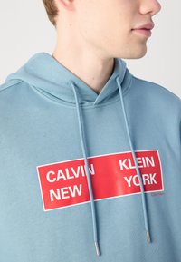 Šviesiai mėlynas švarkelis, pagamintas iš minkštos medžiagos, su ryškiu raudonu stačiakampiu grafiku, kuriame balta užrašas "CALVIN NEW KLEIN YORK" ir sidabriniai aksesuarai ant raištelių.