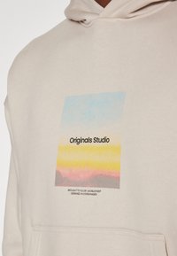 Beige hoodie met een voorkepers, voorzien van een grafisch ontwerp in pastelkleuren (blauw, geel, roze) en de tekst "Originals Studio."