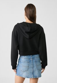 Sudadera negra corta con capucha y mangas largas combinada con una mini falda vaquera azul claro con bolsillos traseros y cierre de cremallera.