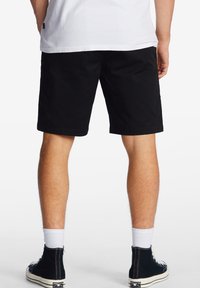 Pantaloni corti neri in cotone con taglio dritto, tasche laterali e una texture liscia, indossati con sneakers alte nere e calzini bianchi.