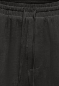 Gros plan sur un pantalon en tissu noir avec une ceinture élastique et deux longs cordons pendants au centre.