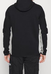 Persona con sudadera negra y paneles laterales grises, y pantalones cargo negros, vista desde atrás sobre un fondo sencillo.