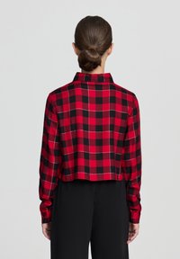 Camicia a quadri rossa e nera con un design cropped, colletto, maniche lunghe e polsini con bottoni. Presenta cuciture bianche nel motivo.