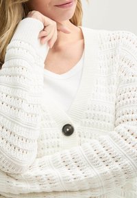 Weißer gestrickter Cardigan mit einem texturierten Muster, gerippten Manschetten und einem einzelnen schwarzen Knopfverschluss. Über einem weißen V-Ausschnitt-Oberteil getragen.