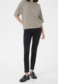 Femme portant un pull en tricot beige à manches courtes, un pantalon slim noir et des mocassins noirs brillants, se tenant contre un fond blanc.