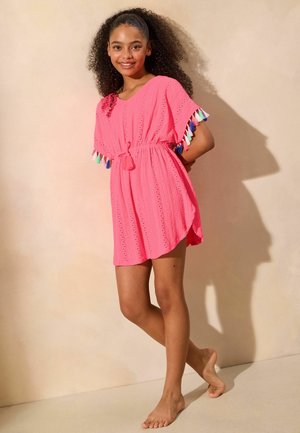 Fată zâmbitoare cu părul creț purtând o rochie roz aprins cu ciucuri multicolori pe mâneci, stând desculță lângă un perete neutru.