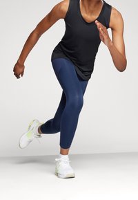 Zwart mouwloos tanktop, marineblauwe leggings en witte sportschoenen met gele accenten. De figuur bevindt zich in een hardlooppose, die de pasvorm van de kleding benadrukt.