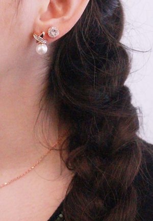 Orecchini in oro rosato con un chiodo circolare decorato da pietre trasparenti e un accento di perla pendente, indossati accanto a un'acconciatura intrecciata.