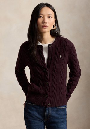 Femme portant un cardigan en maille torsadée violet foncé avec des boutons, sur une chemise blanche et un jean bleu, debout devant un fond neutre.