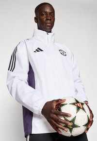Hvid og lilla track jacket med sorte Adidas striber, der har et Manchester United-logo. Modellen holder en hvid fodbold med stjerne mønstre.