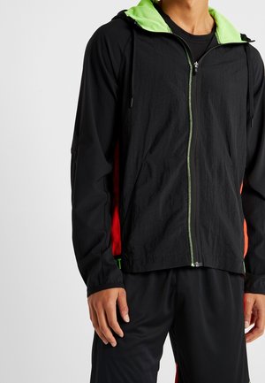 Mann trägt schwarzen Kapuzenpullover mit Reißverschluss, neongrünem Kapuzenfutter und roten Seitenteilen, kombiniert mit schwarzen Hosen.