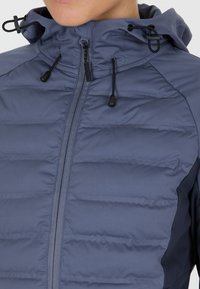 Marineblå puffjakke med lynlås foran, justerbar hætte og kontrasterende sidelommer. Stoffet er glat med kviltet polstring.