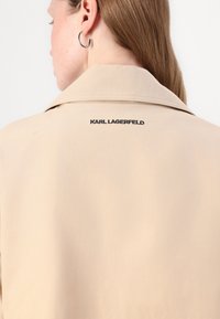 Šviesiai rudas paltas su plačiu apykakle ir matomomis siūlėmis. Nugaroje yra juodos spalvos "KARL LAGERFELD" logotipas. Lygus, lengvas audinys.