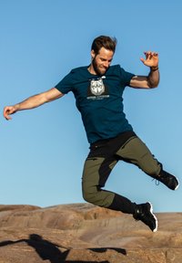 Blå T-shirt med en varg grafik, olivgröna och svarta byxor samt svarta sneakers, fångad mitt i ett språng mot en klar himmel.
