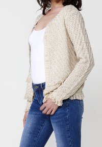 Beżowy dzianinowy kardigan z fakturowanym wzorem i frędzlami. Noszony na białej bluzce z niebieskimi dżinsami. Otwarty przód.