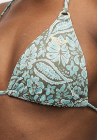 Εκ των κοντών ενός τοπ μπικίνι με φυτικά μοτίβα σε πετρόλ και λευκό χρώμα πάνω σε καστανό δέρμα με μικρό λογότυπο στο πάνω δεξιό τρίγωνο.