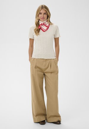 Femeie purtând un tricou bej din tricot cu mâneci scurte, pantaloni largi maro deschis, pantofi maro și un fular alb cu margine roșie legat în jurul gâtului.