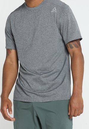 Mężczyzna w szarej koszulce Reebok z krótkim rękawem i zielonych szortach, z tatuażami na przedramionach, stojący na jasnym, jednolitym tle.