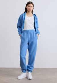 Μπλε βαμβακερό φούτερ με κορδόνι, συνδυασμένο με αντίστοιχο μπλε παντελόνι φόρμας με ελαστική μέση και μανσέτες. Λευκά αθλητικά παπούτσια ολοκληρώνουν την εμφάνιση.