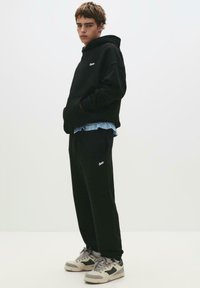 Black hoodie with front pocket, white logo on chest, paired with black sweatpants and gray sneakers, layered over a blue-striped shirt.