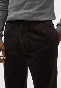 Pantaloni in corduroy di colore marrone scuro. Tessuto a coste verticali con vita elastica e chiusura a bottone. Camicia a maniche lunghe grigia visibile sopra.