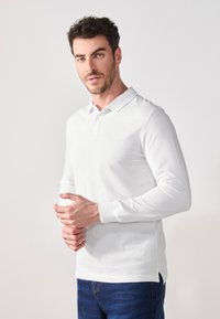 Bílá polo košile s dlouhým rukávem a klasickým límcem, s přímým lemem a bočními rozparky, nošená s modrými džíny. Hladká bavlněná textura.