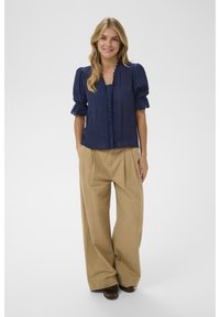 Žena s dlhými blond vlasmi, oblečená v námornícky modrej blúzke s nafúknutými rukávmi a béžových širokých nohaviciach, usmievajúca sa s rukami v vreckách.