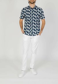 Camicia a maniche corte con motivo geometrico blu e bianco, abbinata a pantaloni bianchi e sneakers bianche; il tessuto sembra liscio e leggero.