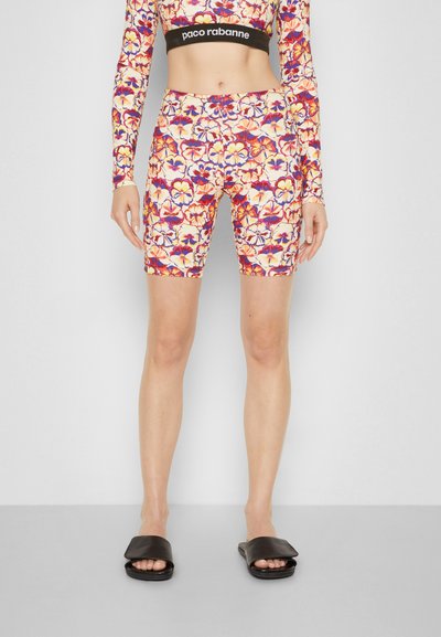 Calções de bicicleta feitos de um tecido elástico com um padrão floral multicolorido em tons de laranja, roxo e amarelo, combinados com chinelos pretos.