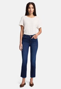 Ciemnoniebieskie dżinsy dzwony z surowym wykończeniem, zestawione z jasnobeżowym t-shirtem z okrągłym dekoltem. Modelka ma warkocze i nosi buty w print.