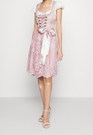 Femme portant une robe en dentelle rose longueur genou avec des manches bouffantes, un nœud en satin à la taille, et des sandales blanches à talons hauts.