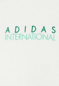 Vit bomullstyg med grön broderad text: "ADIDAS" i versaler och "INTERNATIONAL" under, med varierande nyanser av grönt.