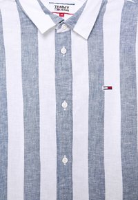 Knappad skjorta i medelstorlek med vertikala vita och grå ränder, med en Tommy Jeans etikett och en liten broderad logotyp på bröstet.