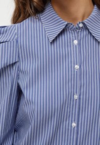 Närbild av en person som bär en blå och vit vertikalt randig skorta med knapp och spetsig krage.