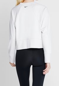 Sweatshirt blanc court avec manches raglan longues, arborant un petit logo Nike noir au dos. Texture douce et lisse ; associé à un legging noir.