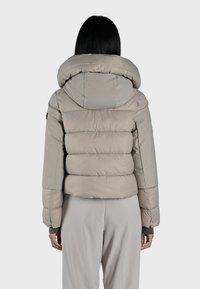 Giacca imbottita beige con un grande cappuccio arrotondato, design trapuntato e polsini elasticizzati. Texture liscia e vestibilità leggermente accorciata.