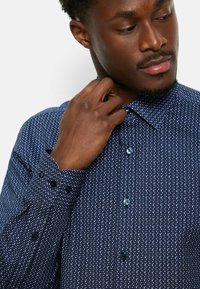 Blaue gemusterte Bluse mit kleinen geometrischen Designs, Knopfleiste und langen Ärmeln mit Knopfmanschetten. Weiche Stofftextur.