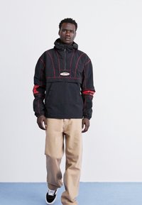 Chaqueta con capucha negra y costuras rojas, un bolsillo frontal y parche con logo; combinada con pantalones beige y zapatillas en blanco y negro.