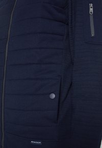 Chaqueta de navy con textura acolchada en el frente, presenta un bolsillo con cremalleras. Fabricada con una tela suave y detalles acanalados. Logo en el dobladillo.