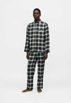 Uomo in piedi a piedi nudi che indossa una camicia a quadri nera, bianca e verde a maniche lunghe e pantaloni a quadri abbinati su uno sfondo uniforme.