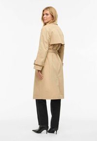 Trench beige avec taille ceinturée, design à double boutonnage, manches longues, épaulettes et une fente au dos pour faciliter le mouvement.